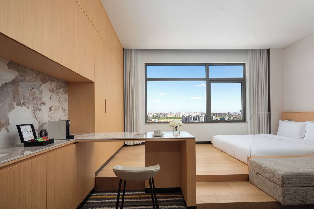 A modern hotel room with a desk, a bed, and a large window offering a city view.