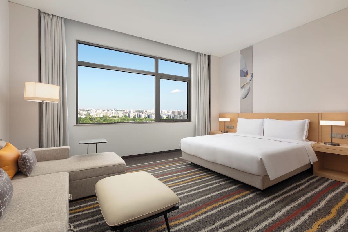 A hotel room with a large window, a bed, a sofa, a small table, and a lamp.