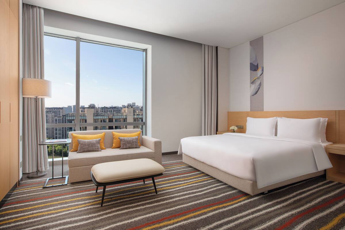 A modern hotel room with a large window, a sofa with cushions, a bed with white linens, and a striped carpet.