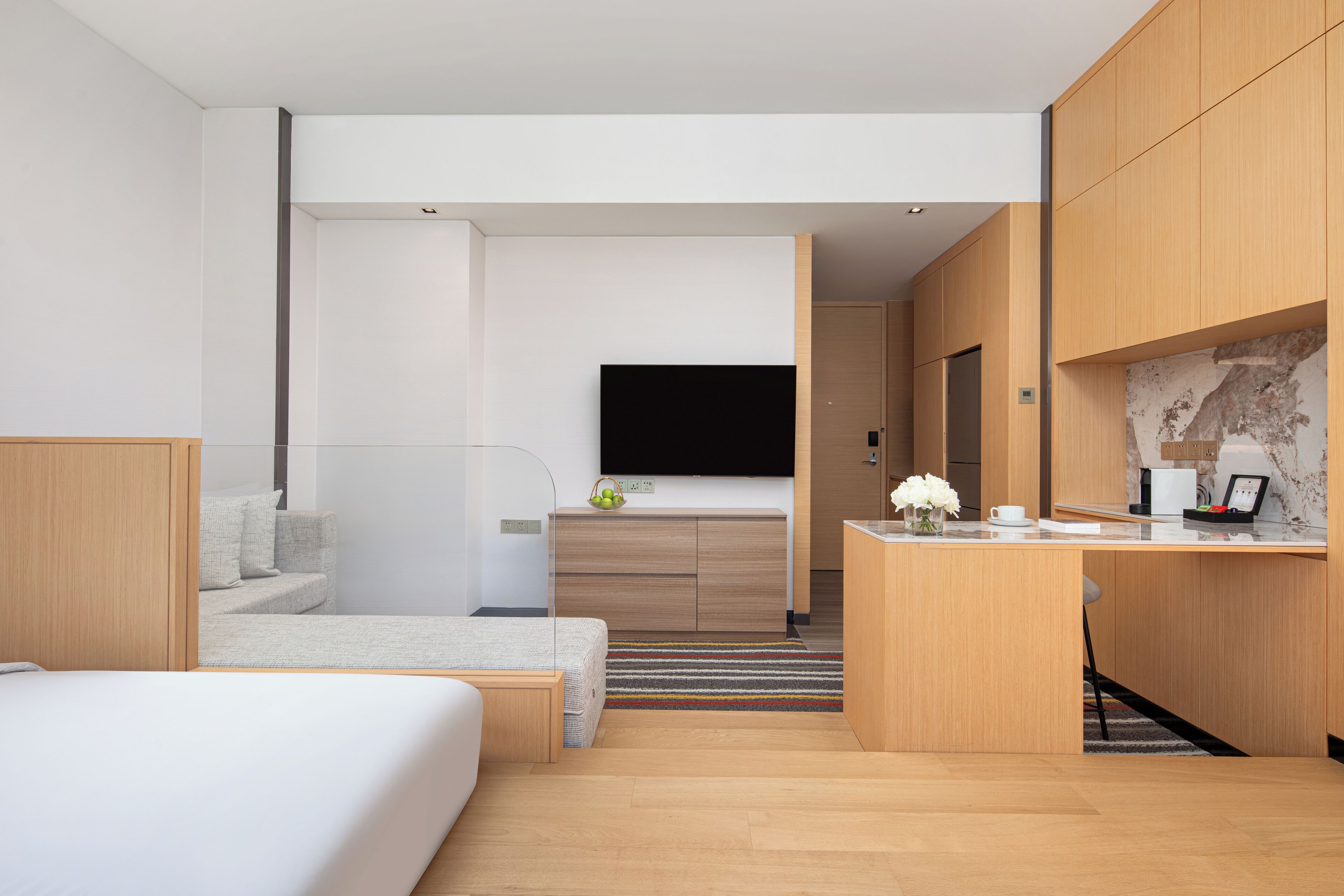 A modern hotel room with a bed, a small seating area, a kitchenette, and a flat-screen TV.