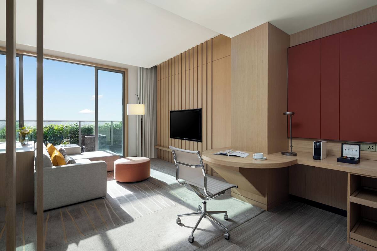 A modern hotel room with a large window, a flat-screen TV, a desk with a coffee maker, a comfortable sofa, and a red cabinet.