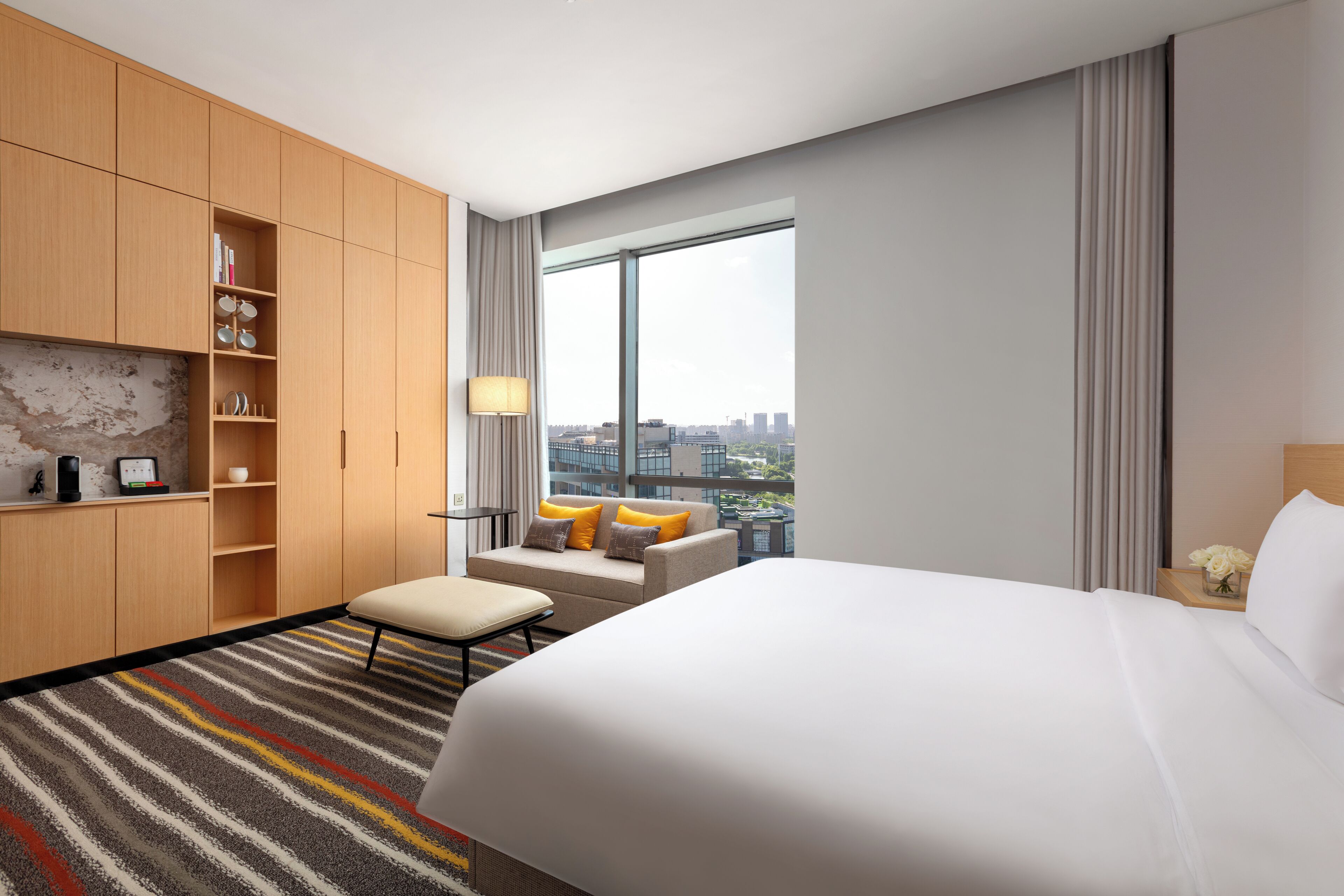 A hotel room with a large bed, a sofa, a small table with a lamp, and a view of the city.