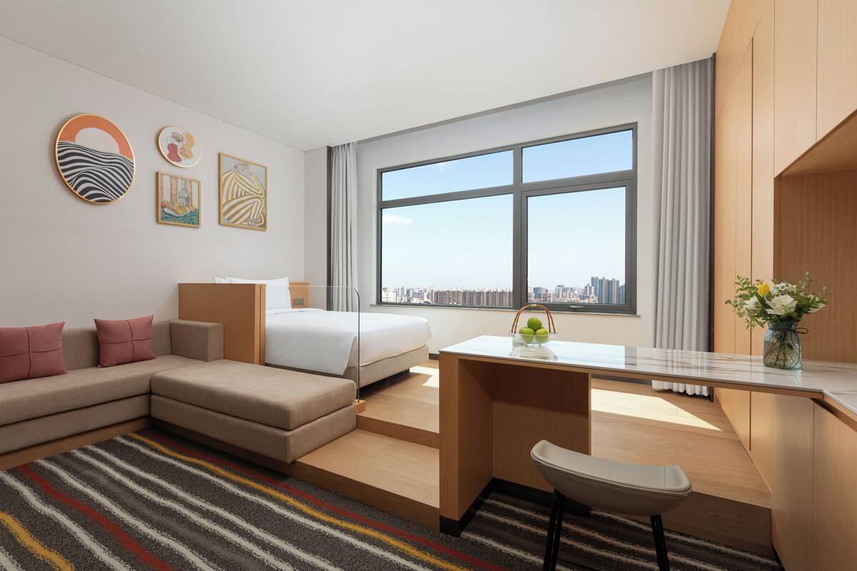 A modern hotel room with a sofa, bed, desk, and large window offering a city view.