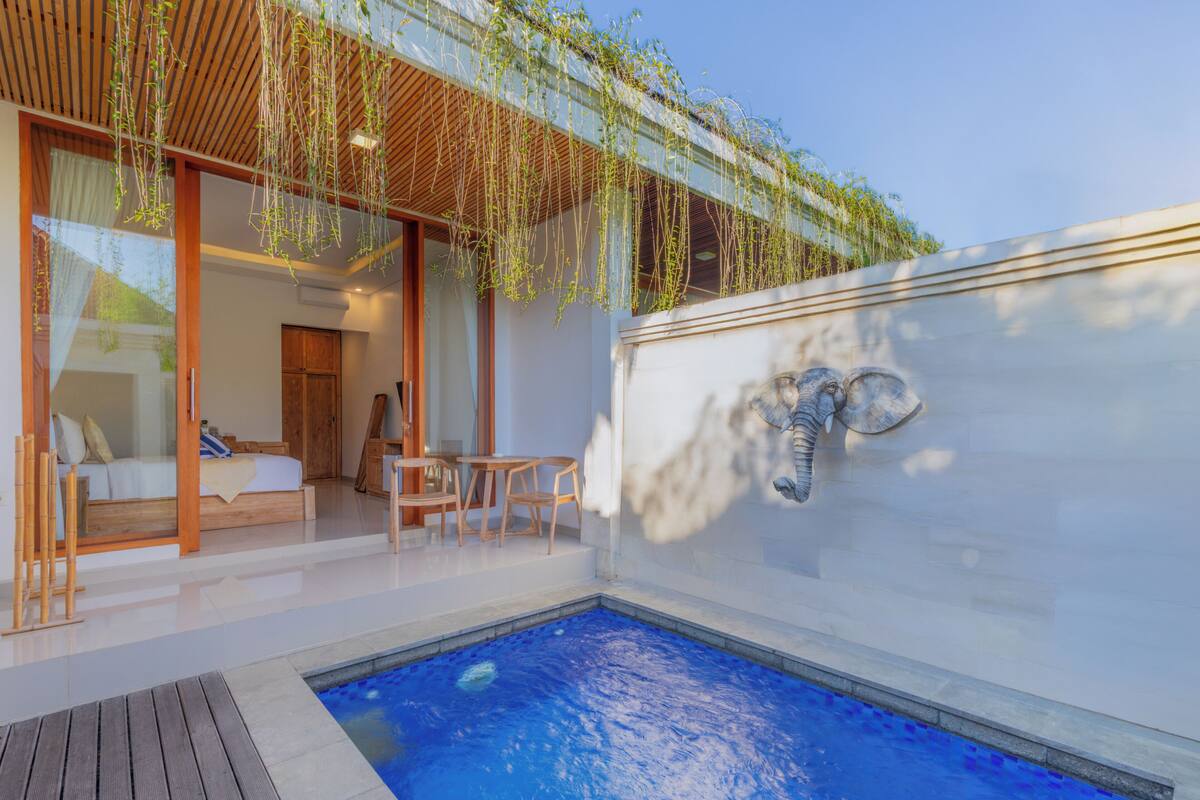 A modern outdoor pool area with a wooden deck, a glass-enclosed room with a bed, and a stone wall with a shell design.