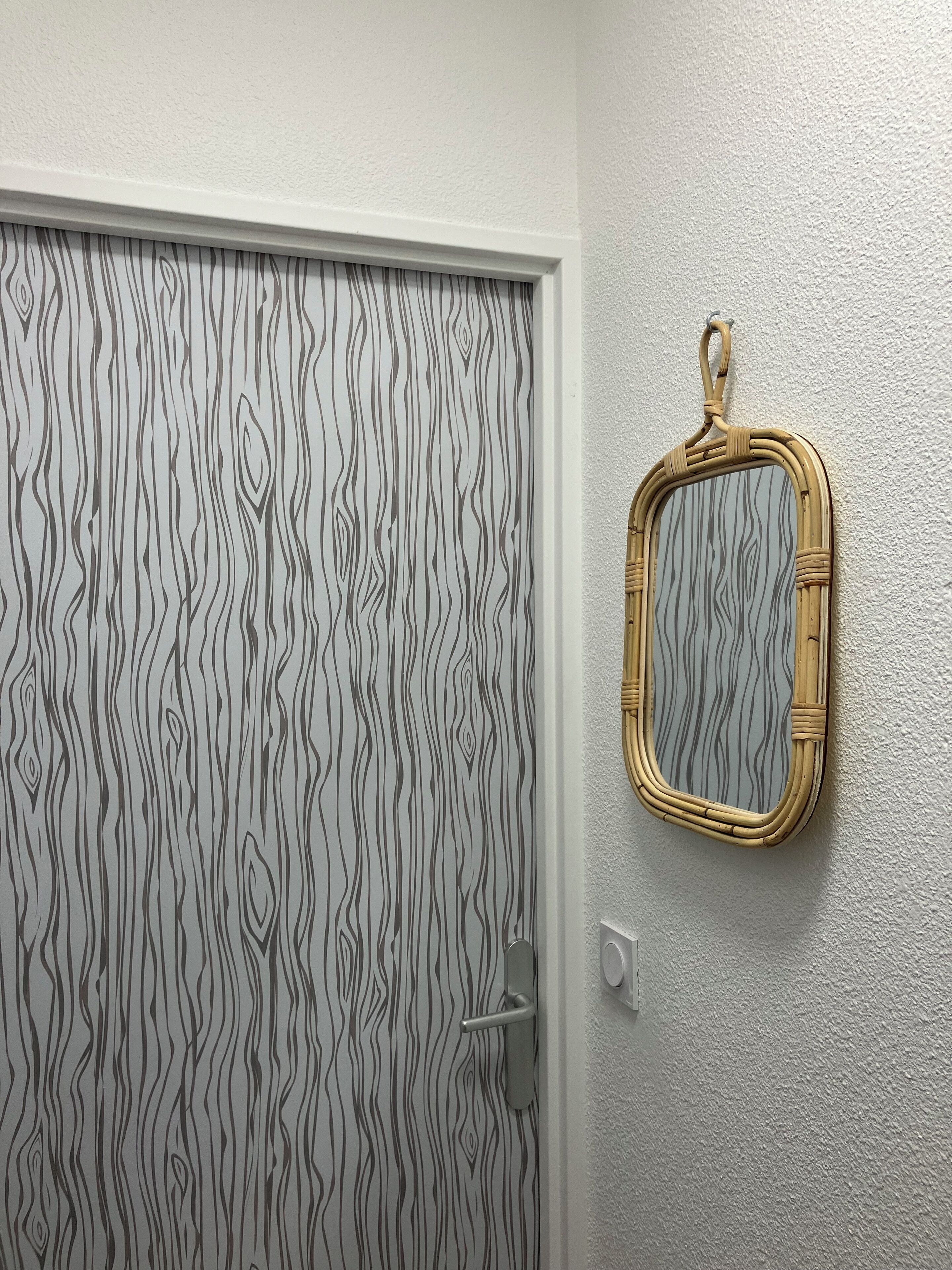 A door with a wavy wood pattern, a mirror with a woven frame, and a light switch on the wall.