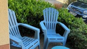 Two blue Adirondack chairs and a small round table on a gravel surface, with a parking lot and greenery in the background.