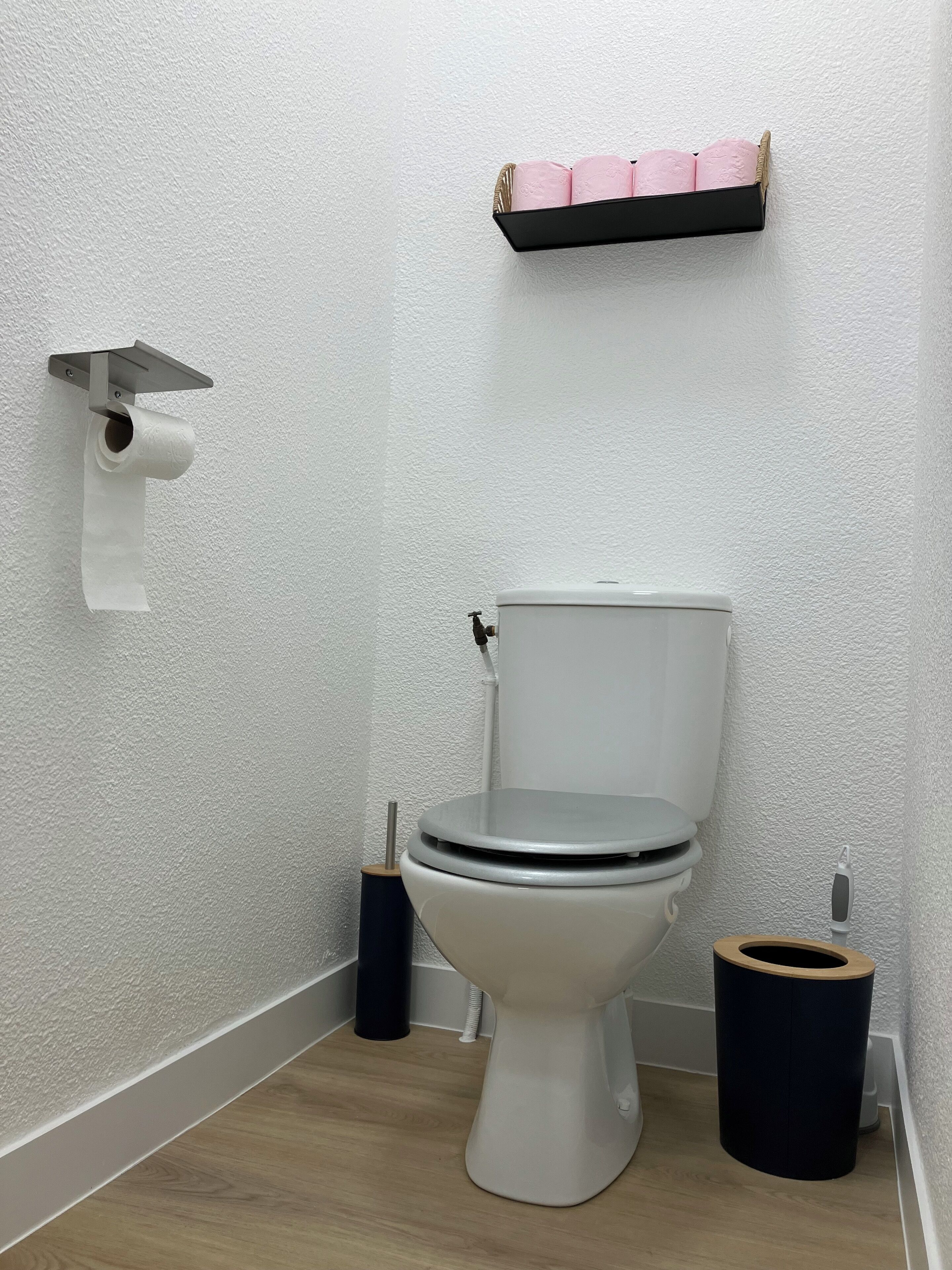 Een badkamer met een toilet, toiletrolhouder, prullenbak en een plank met roze spullen.