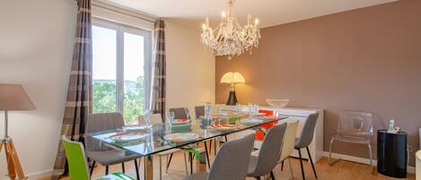 A dining area with a glass table, grey and green chairs, a chandelier, and a view through large windows.