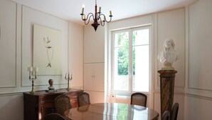 Ein Esszimmer mit rundem Tisch, sechs Stühlen, einer Chandelier, einer Büste auf einem Sockel, einem Kerzenleuchter und einem Spiegel mit einer Pflanze.