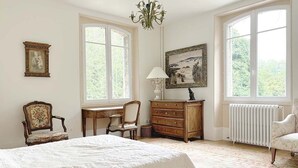 Ein Schlafzimmer mit einem großen Bett, zwei Stühlen, einem Schminktisch, einem Schreibtisch, einer Lampe, einem Bild und einer Heizung.