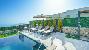 Ein Infinity-Pool mit Liegestühlen und Sonnenschirm, umgeben von einer Steinmauer und Grünpflanzen, mit Blick auf eine schöne Aussicht.