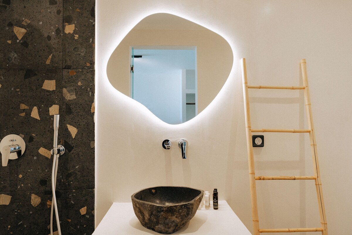 A bathroom with a stone sink, a mirror with a unique shape, a bamboo ladder, and a shower with a modern faucet.
