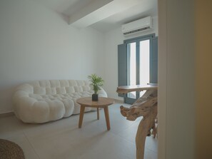 A room with a tufted sofa, a round wooden table with a potted plant, and a rustic wooden coffee table. - Anemos Imerovigli Suites (Santorini)