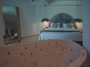A bedroom with a bed, a large mirror, a round table with a bowl of fruit, and a unique ceiling fan. - Anemos Imerovigli Suites (Santorini)