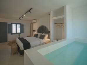 A modern bedroom with a large bed, a hanging light fixture, a window with shutters, and a built-in swimming pool. - Anemos Imerovigli Suites (Santorini)