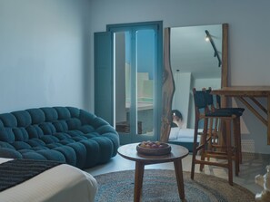 A modern living room with a sofa, a small round table with fruit, and a wooden chair. - Anemos Imerovigli Suites (Santorini)