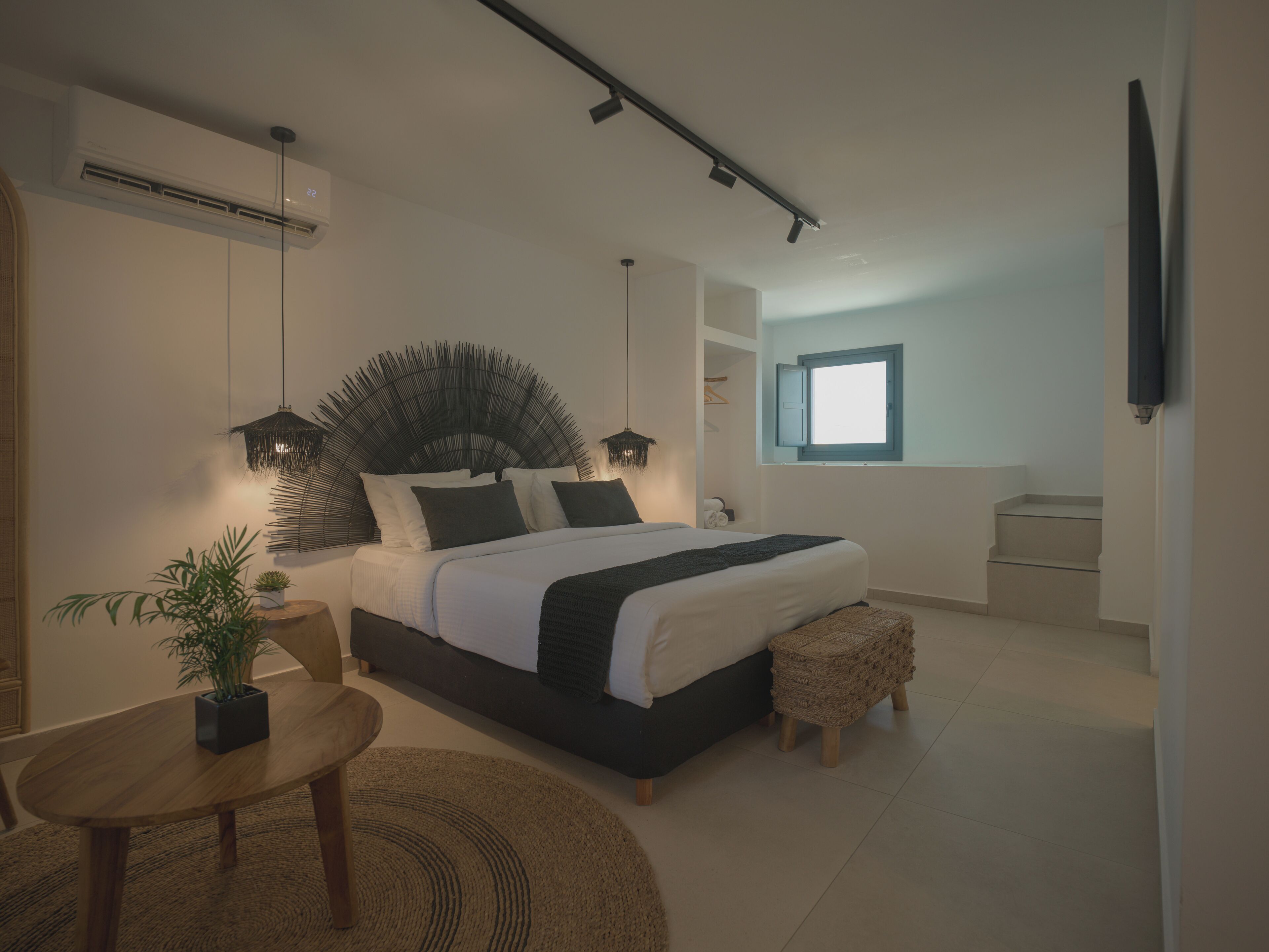A modern bedroom with a large bed, bedside tables, a small table, a potted plant, and a window.