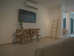 A modern living room with a large flat-screen TV mounted on the wall, a wooden console table, a potted plant, and a ladder leaning against the wall. - Anemos Imerovigli Suites (Santorini)
