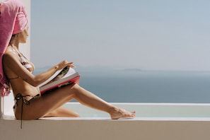 A person sitting by a window with a view of the sea, wearing a towel and reading a book. - Anemos Imerovigli Suites (Santorini)