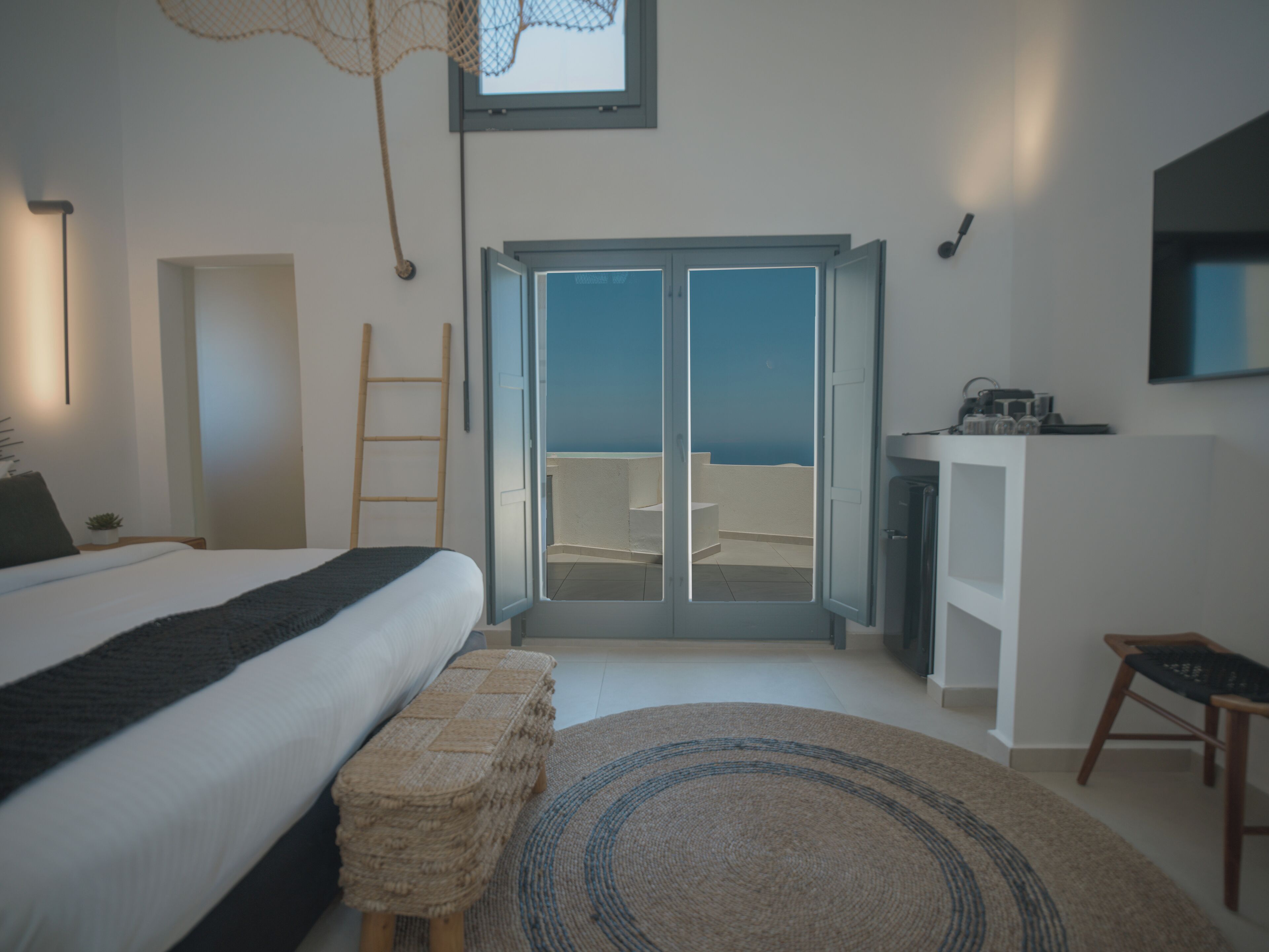 A bedroom with a bed, a bench, a desk, and a view of the sea through a sliding door.