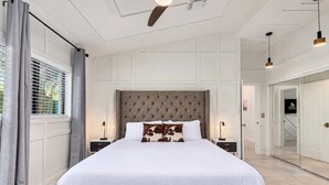 Une chambre avec un grand lit, une tête de lit capitonnée, des tables de chevet avec des lampes et une vue sur un couloir avec une porte.