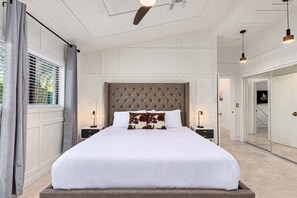 Ein Schlafzimmer mit einem großen Bett, einem gepolsterten Kopfteil, Nachttischen mit Lampen und Blick auf einen Flur mit einer Tür.