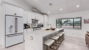 Uma cozinha moderna com armários brancos, uma ilha grande com banquetas, eletrodomésticos de aço inoxidável e uma janela com vista para o exterior.