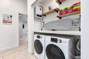 Une buanderie équipée de deux machines à laver à chargement frontal, d’un radiateur et d’étagères où sont rangées des serviettes colorées, ainsi qu’un tableau à repasser.