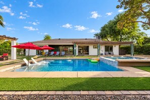 A residential swimming pool with lounge chairs and umbrellas, surrounded by a well-maintained lawn and a house with a patio.