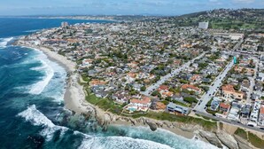 Vista aérea de un pueblo costero con playa, zonas residenciales y un paisaje montañoso al fondo.
