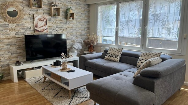A modern living room with a stone wall, a flat-screen TV, a grey sofa, and a large window with a view of trees.