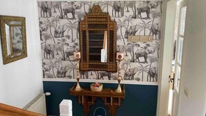 A room with a patterned wallpaper depicting elephants and palm trees, a wooden console table with a mirror and speakers, and a white radiator.