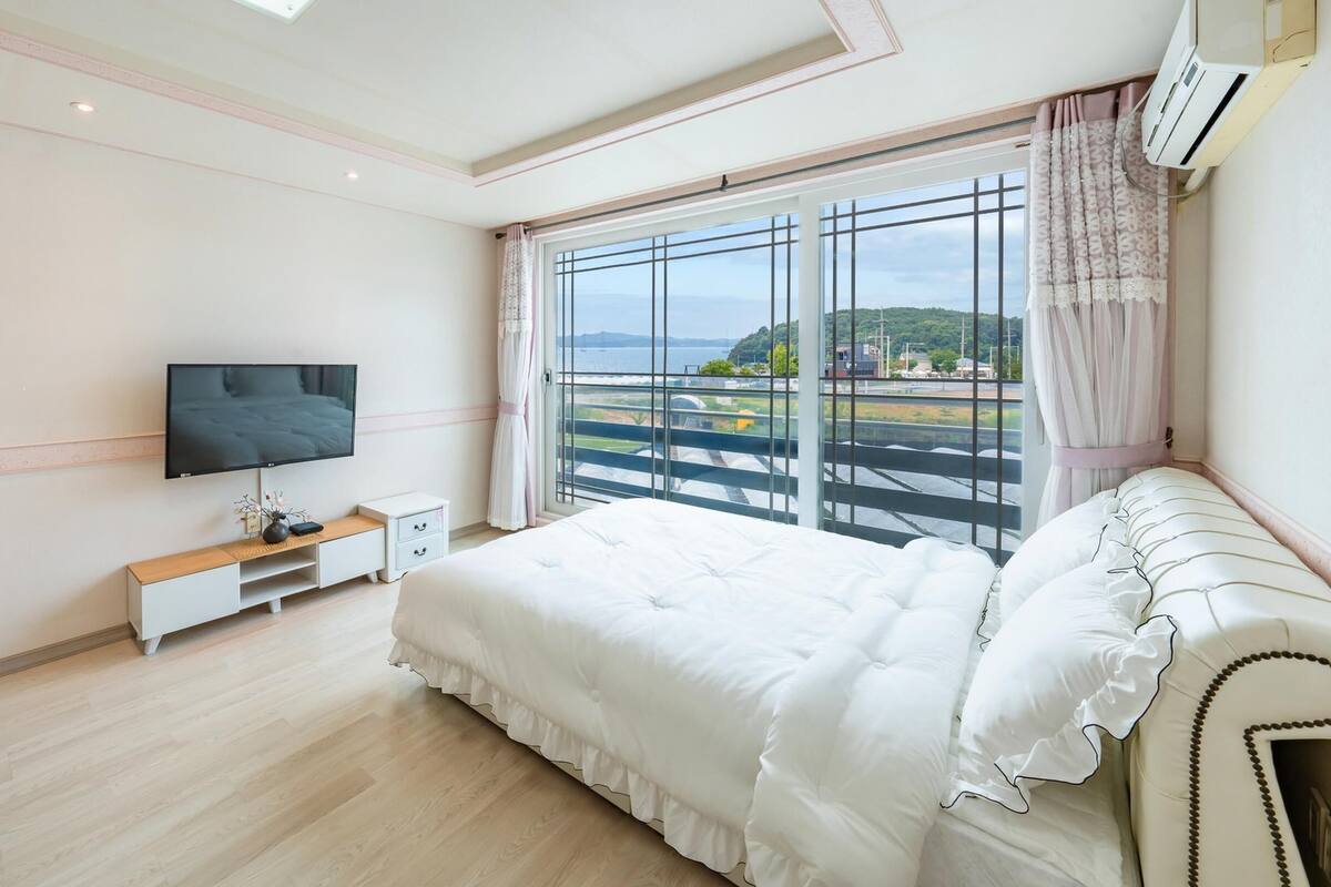 A bedroom with a large bed, a TV mounted on the wall, a bedside table, and a window with a view of a landscape.