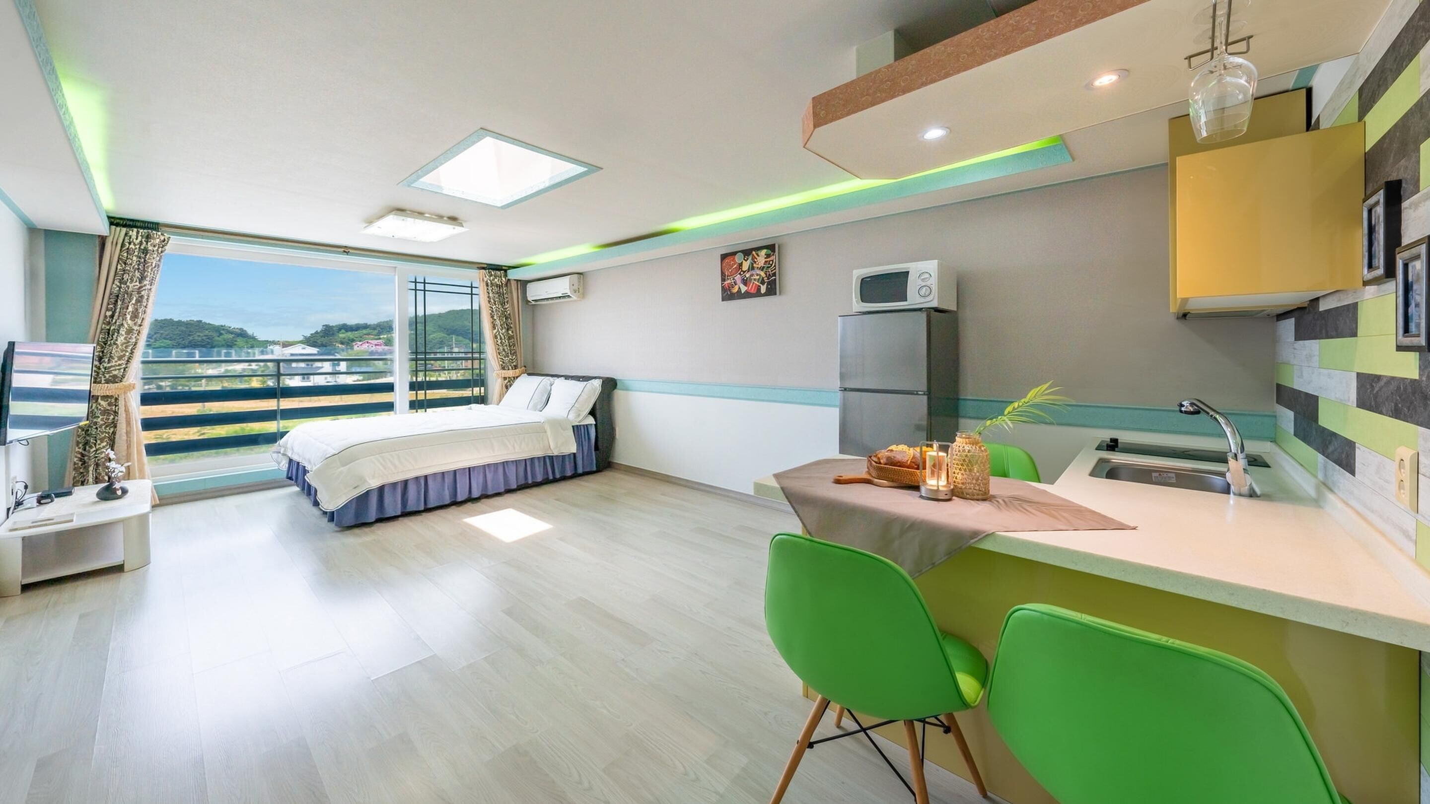 A modern hotel room with a balcony, a bed, a desk, a kitchenette, and a dining area.