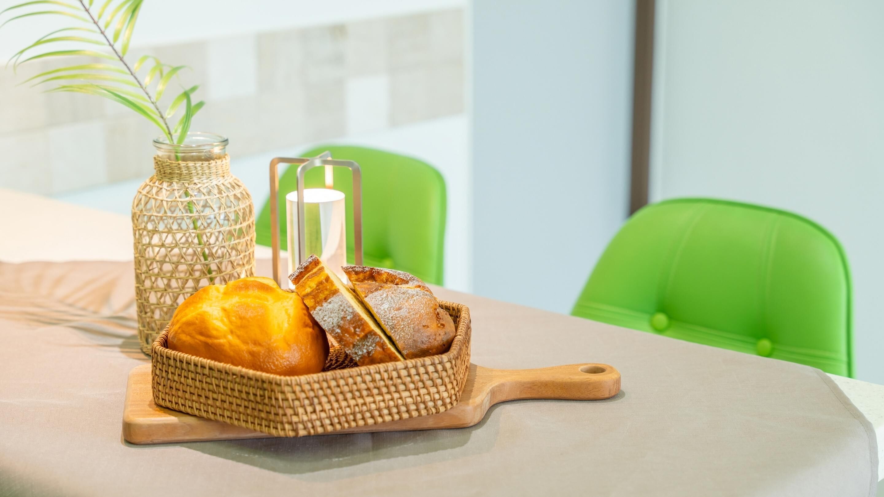 A breakfast setup with bread and a woven basket on a table, a woven vase with a plant, and a green chair in the background.