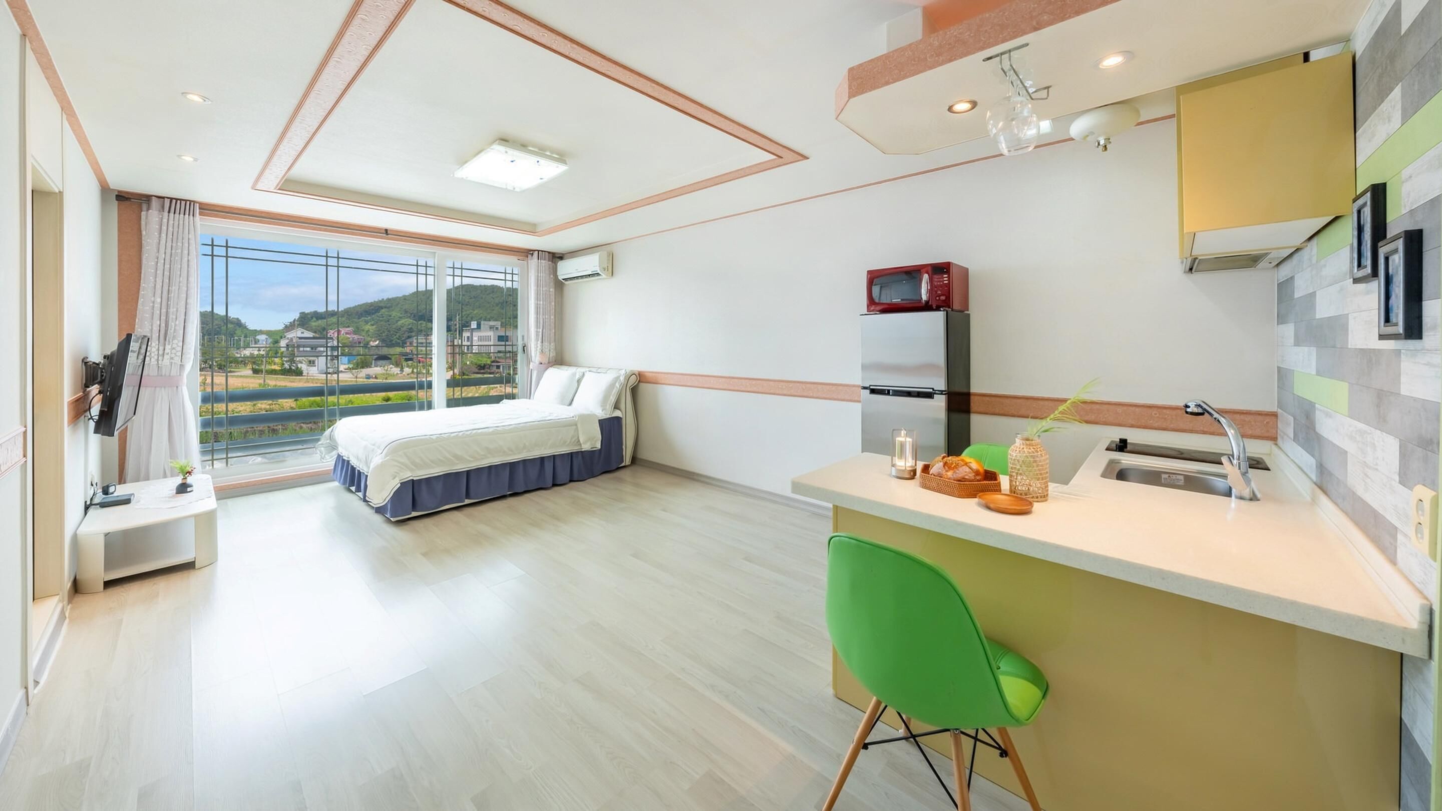Ein modernes Hotelzimmer mit Bett, Schreibtisch, grünem Sessel, Küchenzeile und großem Fenster mit Blick auf die Landschaft.