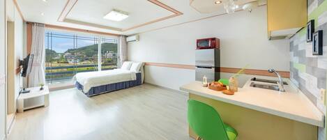 Ein modernes Hotelzimmer mit Bett, Schreibtisch, grünem Sessel, Küchenzeile und großem Fenster mit Blick auf die Landschaft.