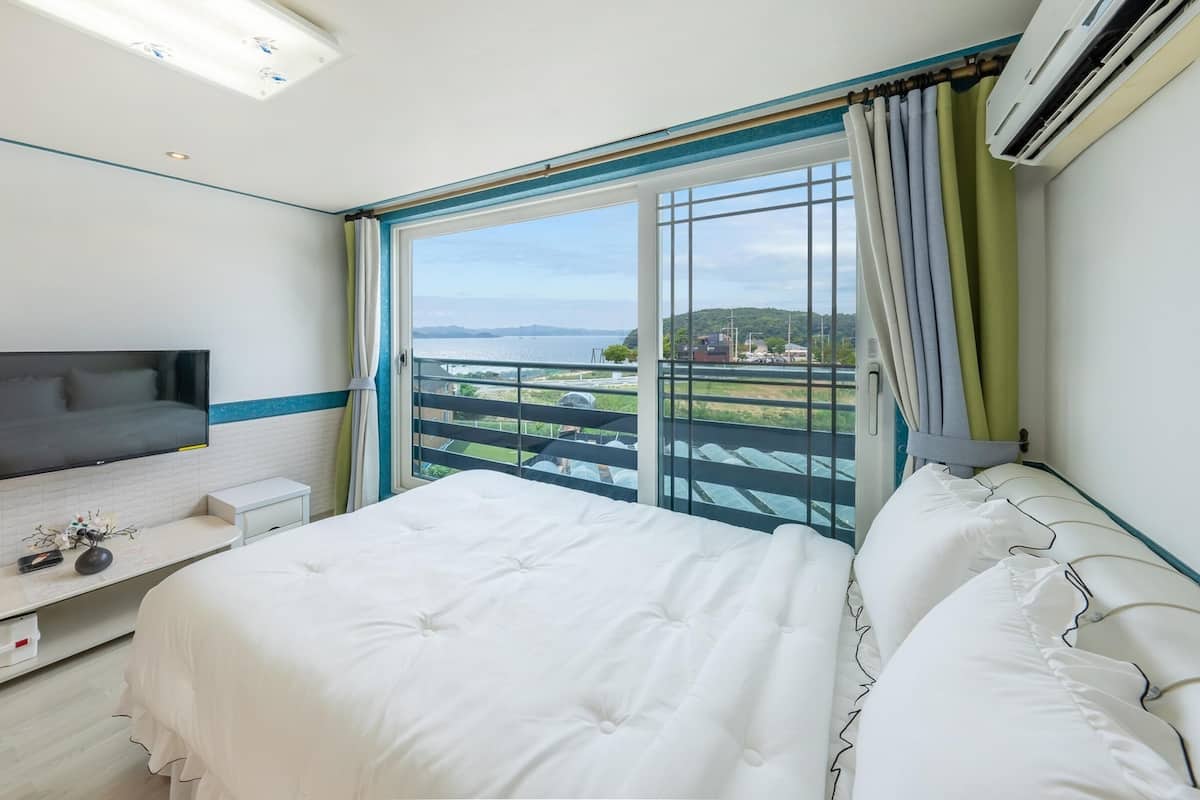 A hotel room with a bed, a TV, a small bedside table, a window with a view of the sea, and an air conditioning unit.