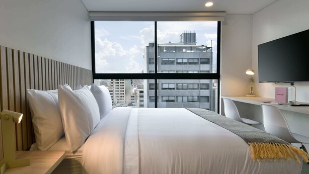 A hotel room with a large bed, a desk with a chair, a TV, and a window with a city view. - w* | Perfect Loft in Santa Barbara (Bogotá)