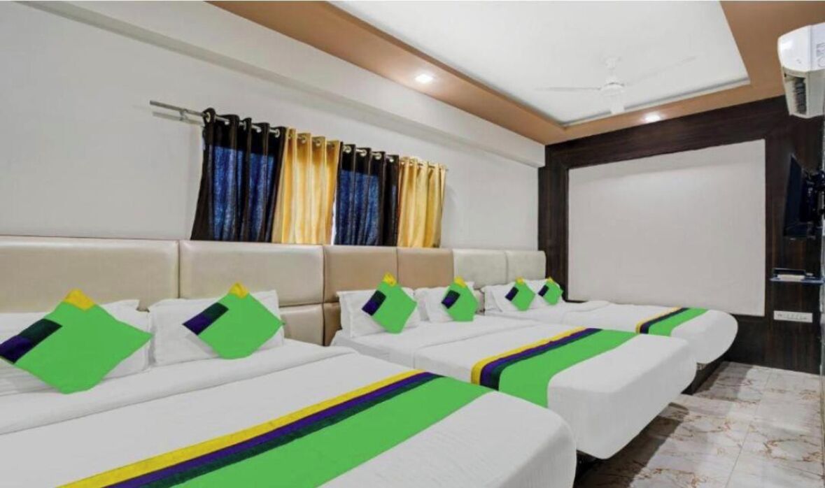 Quarto de hotel com quatro camas de solteiro, cada uma com colcha e almofadas verdes e roxas. Há uma janela grande com cortinas, um ventilador de teto e uma tela de projetor na parede.