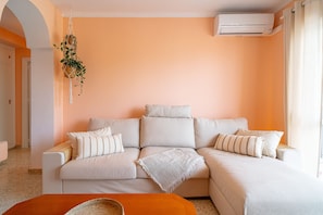 Ein modernes Wohnzimmer mit einem beigen Sofa, einem kleinen orangefarbenen Hocker und einer hängenden Pflanze.