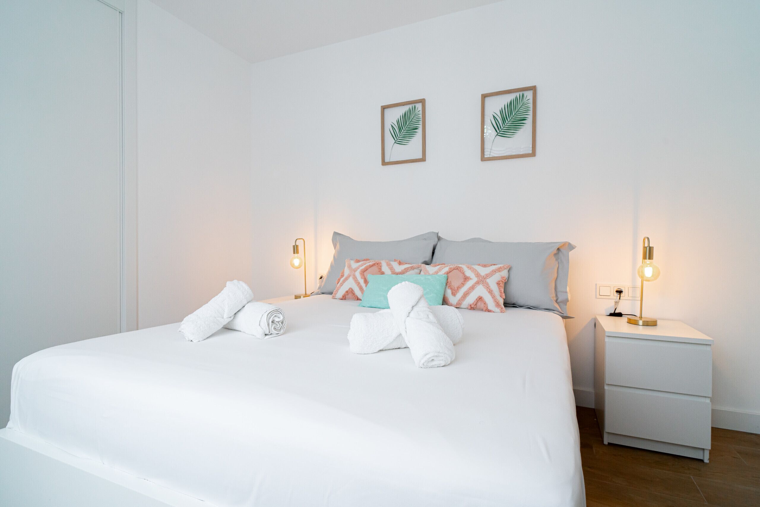 Un lit bien fait, avec du linge de lit blanc et des serviettes pliées, une table de chevet avec une lampe, et deux décorations murales représentant des feuilles, encadrées.