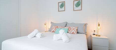 Un lit bien fait, avec du linge de lit blanc et des serviettes pliées, une table de chevet avec une lampe, et deux décorations murales représentant des feuilles, encadrées.