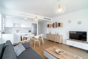 A modern kitchen with white cabinets, a dining area with a wooden table and chairs, a living room with a gray sofa, and a TV on a wooden stand.