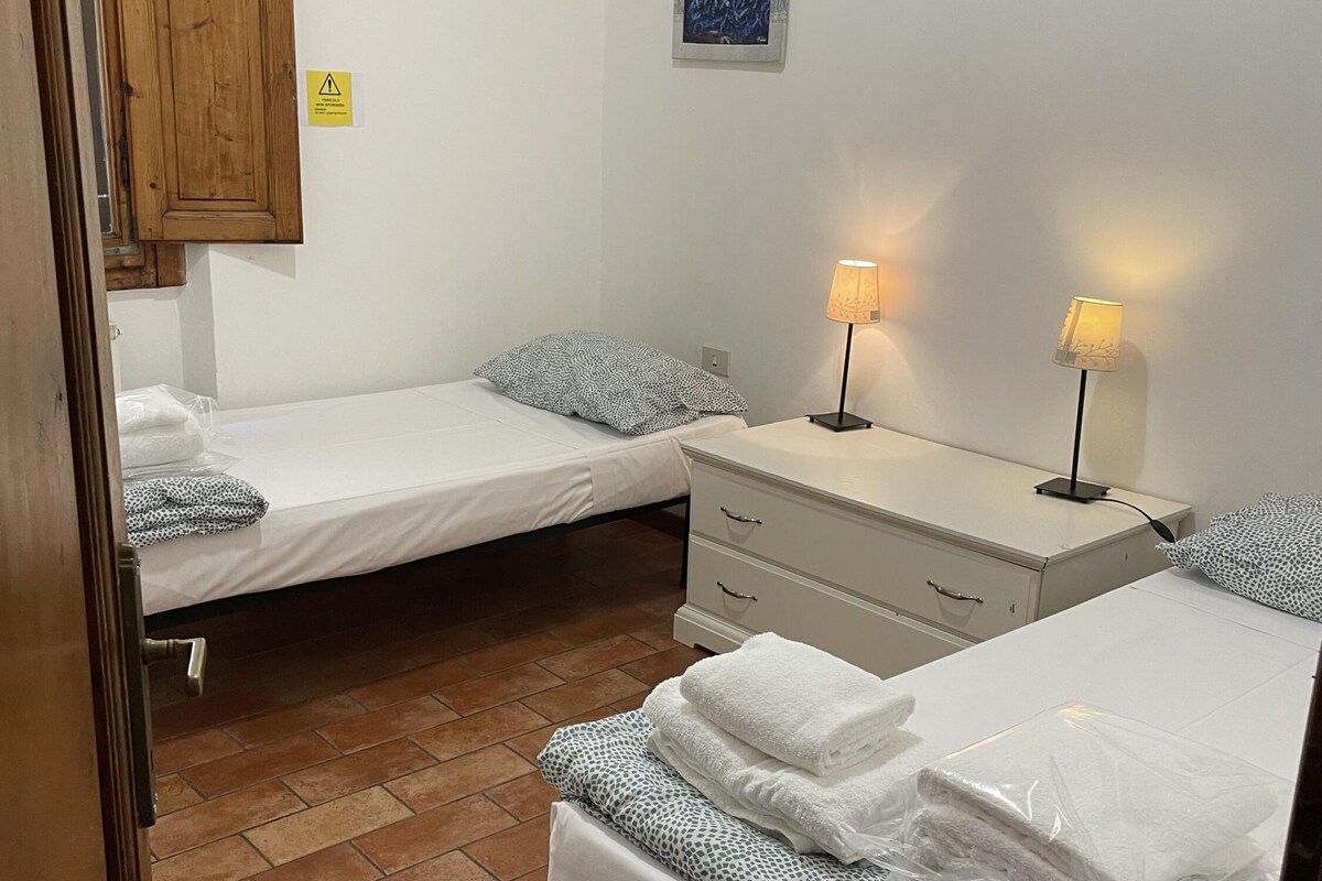 A small, clean room with two single beds, a bedside table with lamps, a wooden wardrobe, and a tiled floor.