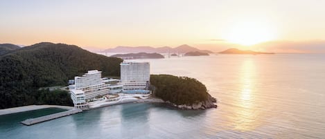 A coastal hotel with a modern design, situated on a small island with a view of the sea and distant mountains.