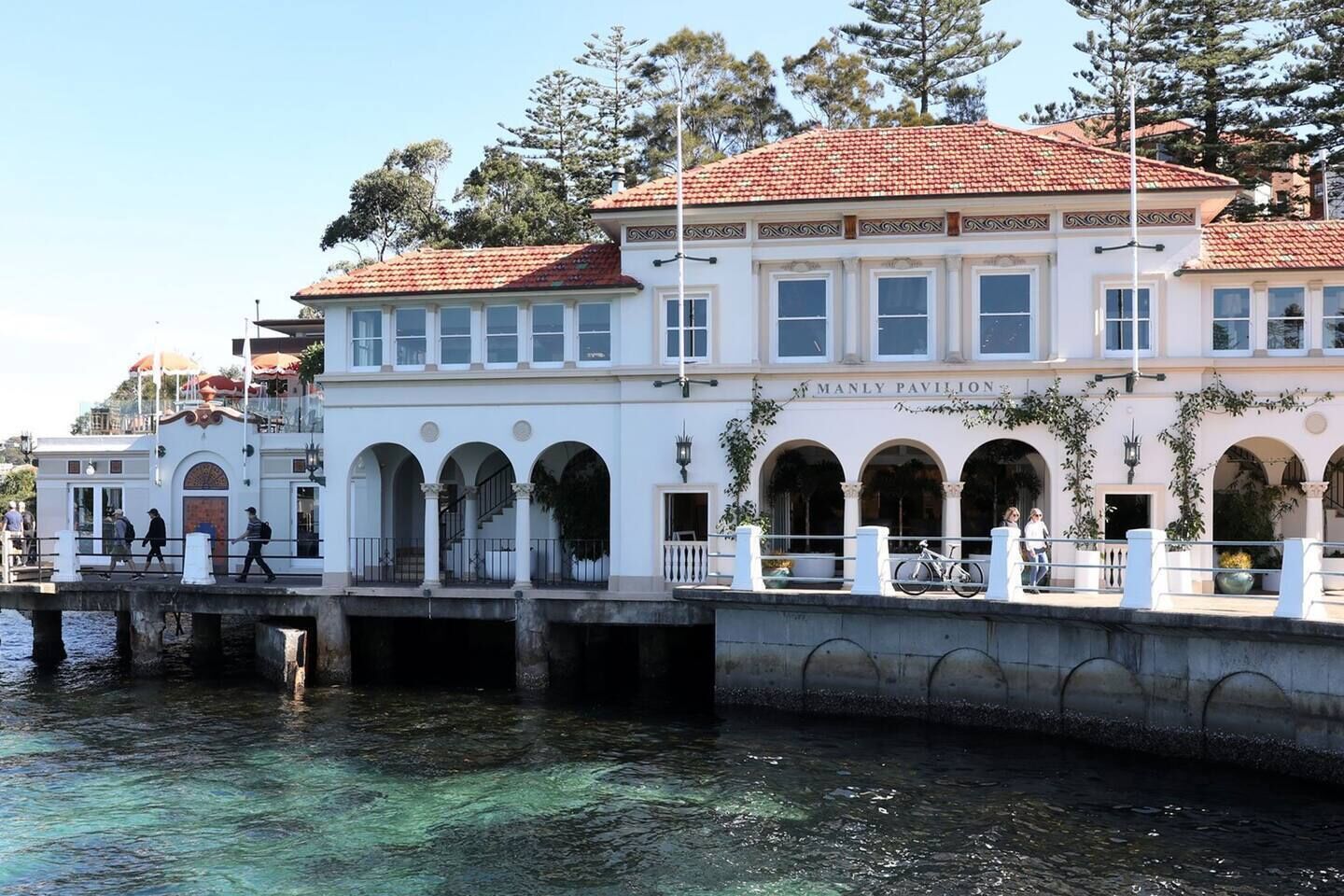 A waterfront building with multiple arches, a balcony, and a sign reading 'Manly Pavilion.'