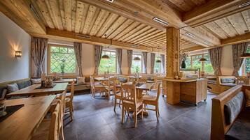 A spacious dining area with wooden tables and chairs, large windows with curtains, and a high wooden ceiling.