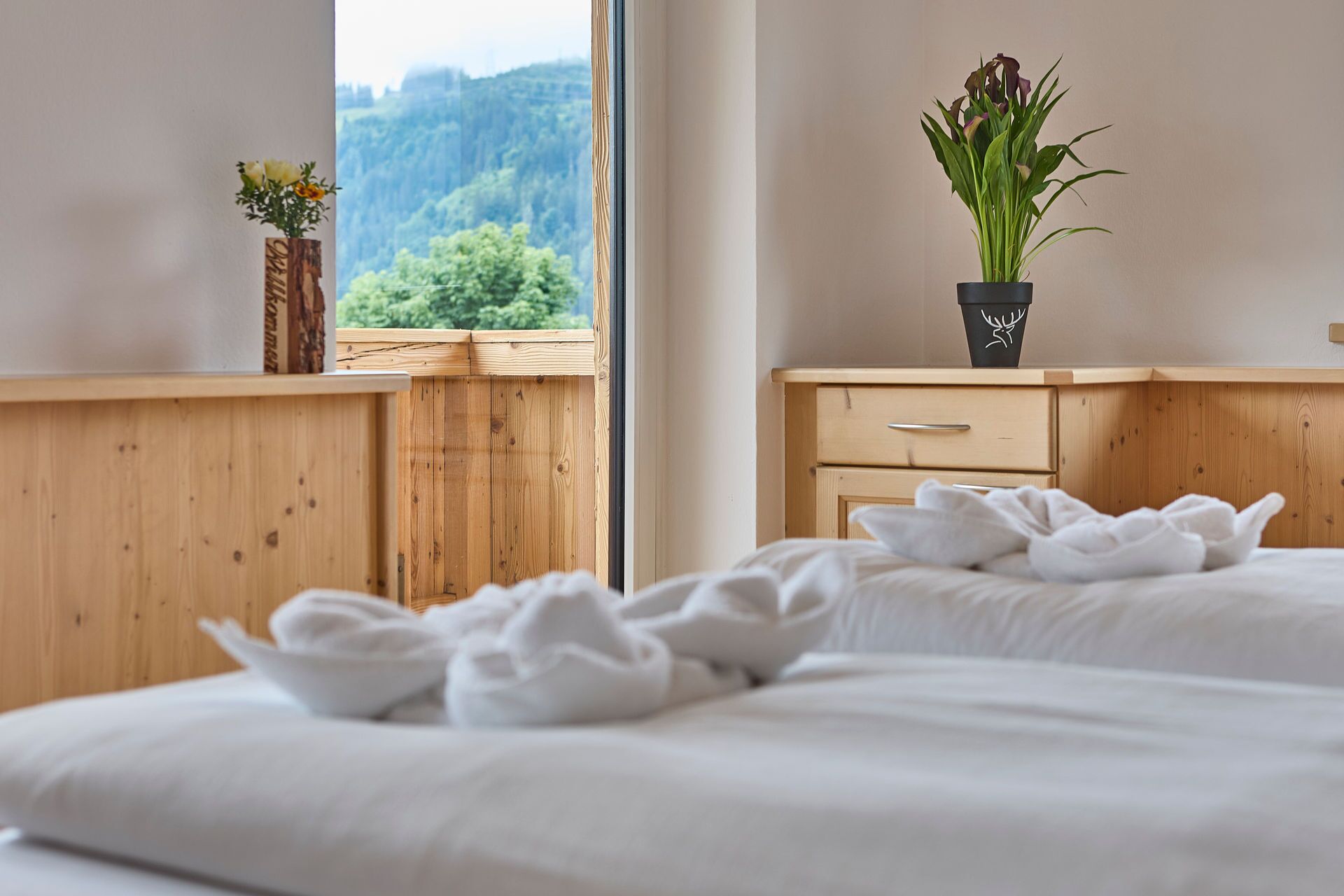 Une chambre à coucher avec un lit en bois, une table de chevet ornée d’un vase de fleurs et une vue sur une forêt, visible par la fenêtre.