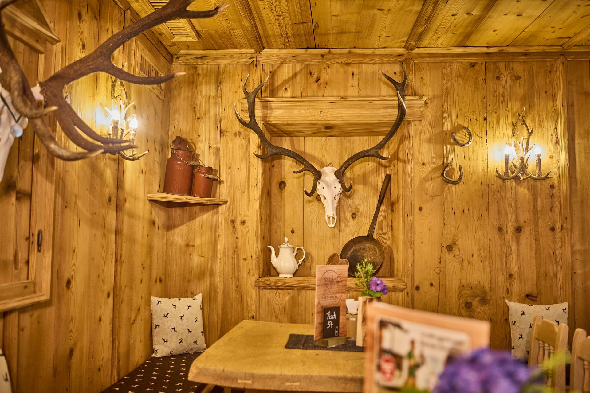 A rustic wooden interior with a table, chairs, a shelf with jars, a skull, and wall-mounted lights.